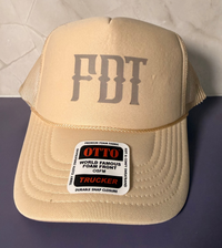 A beige trucker hat with 'FDT' printed on the front, resting on a dark purple surface against a light gray tiled background.