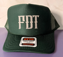 A dark green trucker hat with 'FDT' embroidered in white on the front, resting on a purple surface against a light beige tiled background.