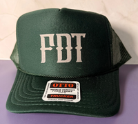 A dark green trucker hat with 'FDT' embroidered in white on the front, resting on a purple surface against a light beige tiled background.