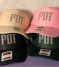 Four FDT trucker hats in beige, pink, black, and green, arranged in a stack on a light marble-patterned surface.